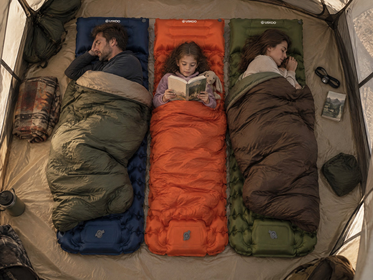 A family of three in a tent at dawn on navy, orange and olive AirFlex pads side by side, the parents asleep, the child reading