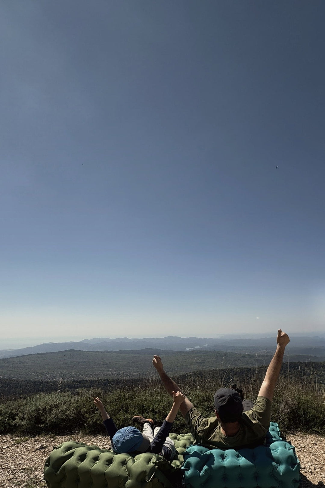 A father and son lying back on two AirFlex pads at a mountain lookout, arms raised toward the valley