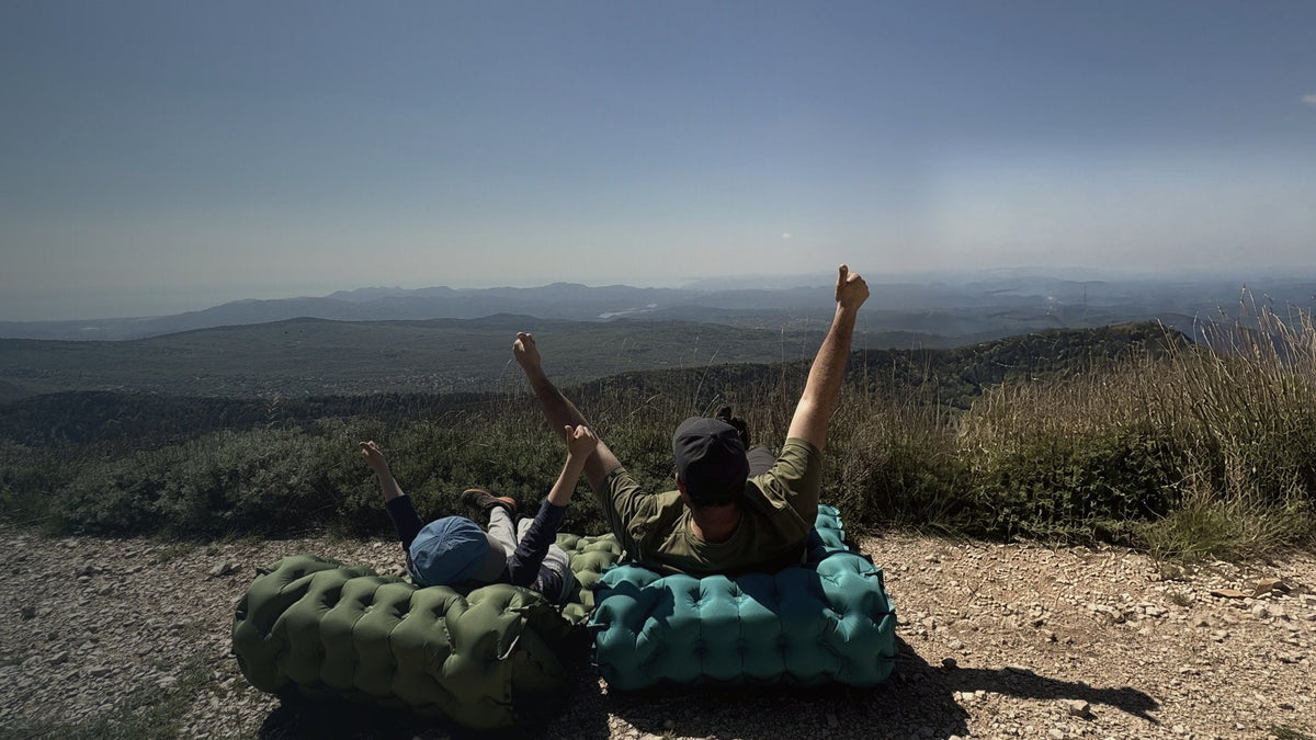 A father and son lying back on two AirFlex pads at a mountain lookout, arms raised toward the valley