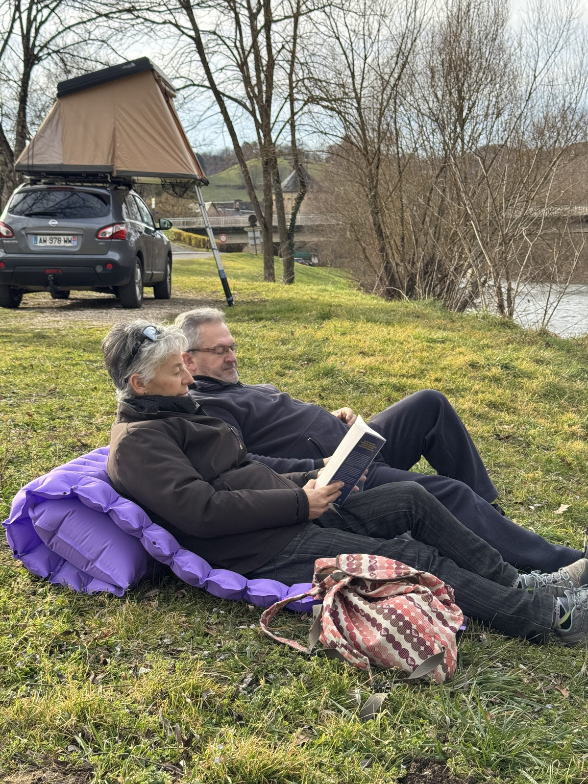 Two campers reading on an AirFlex in lounger mode by a river, rooftop tent behind them